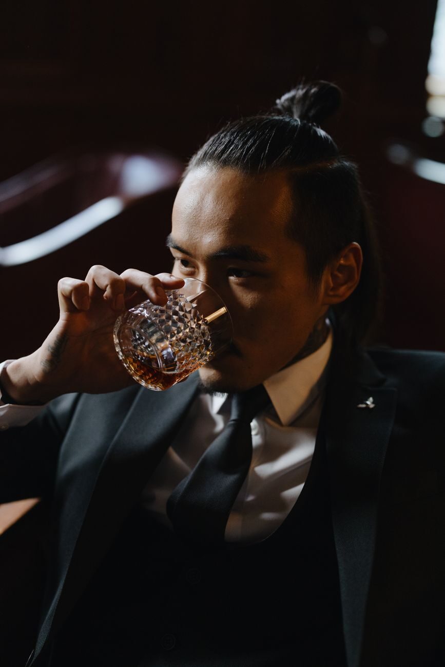 A man in a dark suit sipping an amber cocktail from a cut-crystal glass in warm, low restaurant light